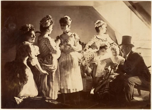 [A Group of Six Costumed Women Posed in Interior with Top Hatted Gentlemen]