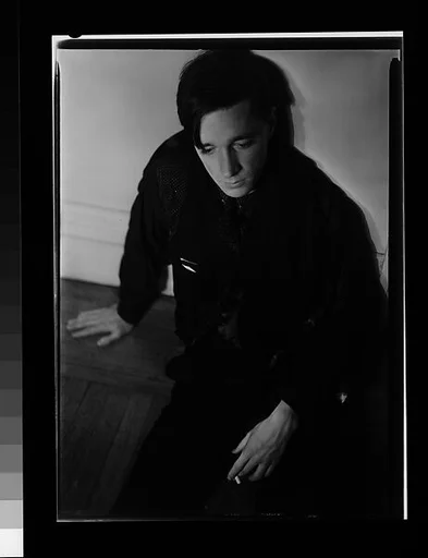 [Self-Portrait Seated on Floor Against Wall with Dark Cloth Around Neck]