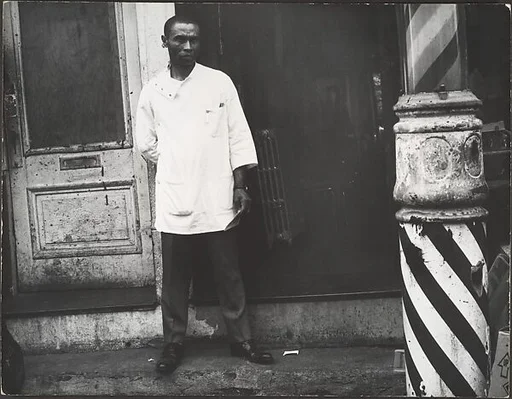 [Barber in White Coat Standing on Street Beside Striped Pole at Entrance to Shop, New York]