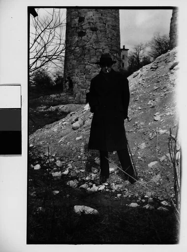 [Hanns Skolle in Overcoat and Hat Before Stone Tower]