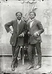 Two dapper young men leaning on a chair