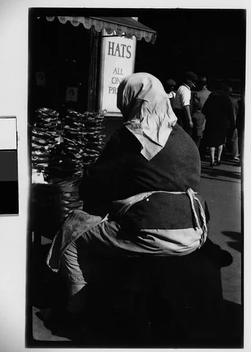 [Street Vendor, From Behind, in Front of Hat Stand, Possibly Delancey Street, New York City]