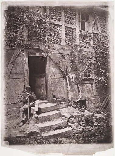 [Man Sitting on Steps of House with Socks Hanging on Nearby Vine to Dry]