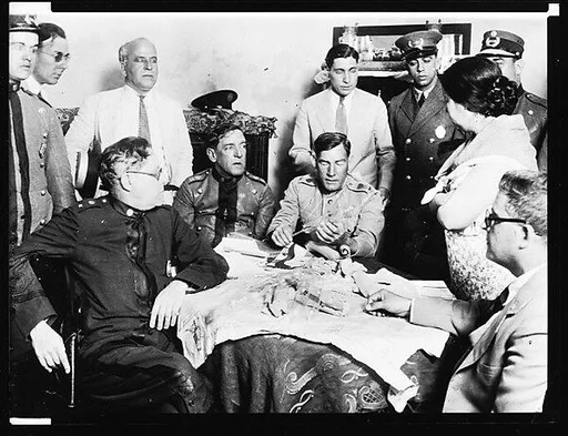[Copy of Newspaper File Photograph: Men and Officers Seated and Standing Around Table]