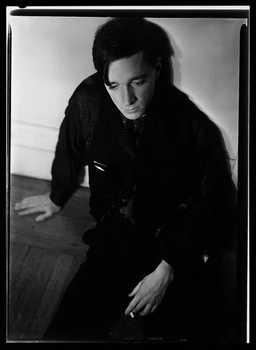 [Self-Portrait Seated on Floor Against Wall with Dark Cloth Around Neck]