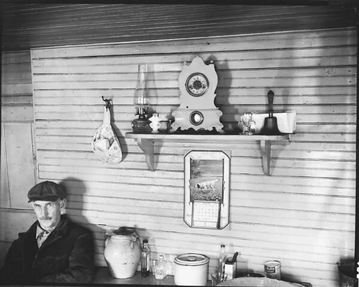 [Man in Cap Seated at Table Below Mantle with Clock, Kerosene Lamp, and Bell, Ossining, New York]