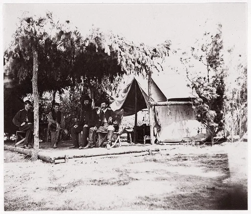[Four men in camp under a lean-to of pine boughs]