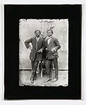 Two dapper young men leaning on a chair