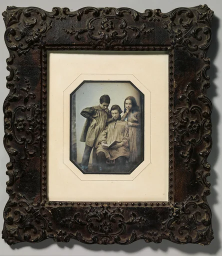 [Three Children, One Seated Holding an Open Book, the Other Two Standing, in Front of a Light Background]